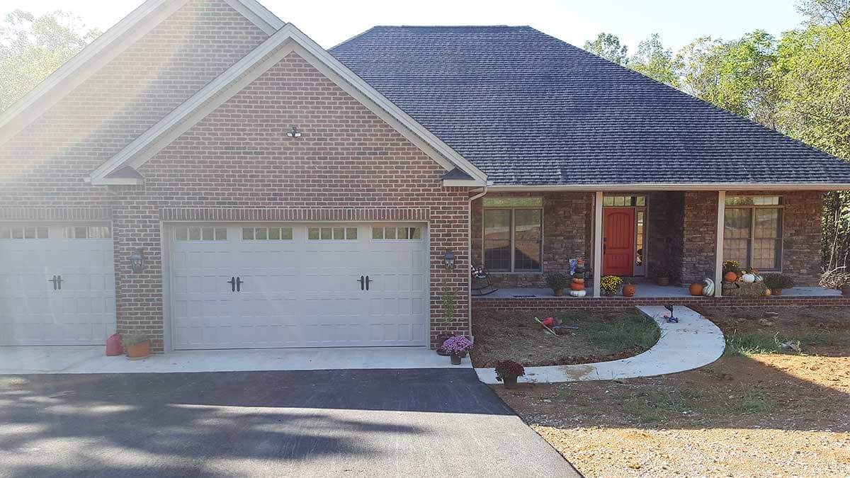 House plan exterior: Two-story brick home with a side-entry two-car garage, gable roof, and covered front porch with a red door.
