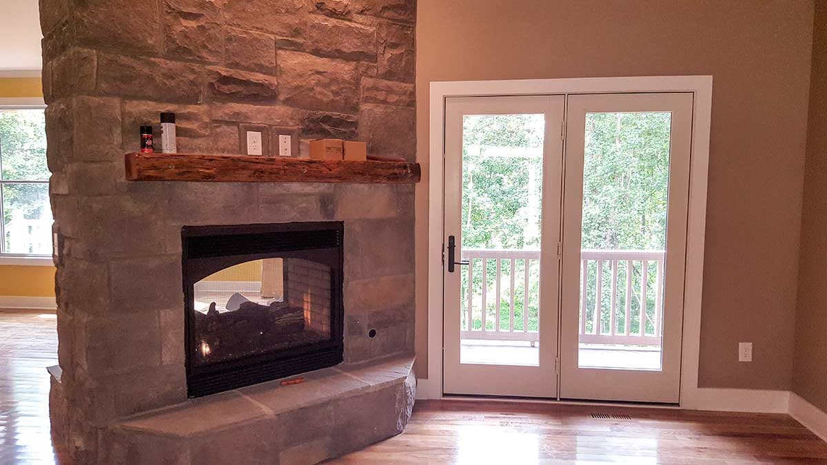 Interior view of a living room with a stone fireplace, wood mantel, and double glass doors leading to an exterior deck.