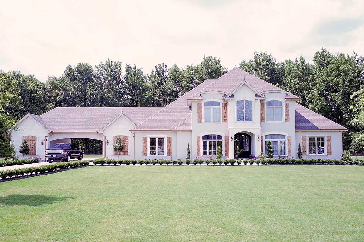 House plan exterior with two stories. Features include a porte-cochere, gable roof, arched windows, and wood shutters. Landscaped lawn.
