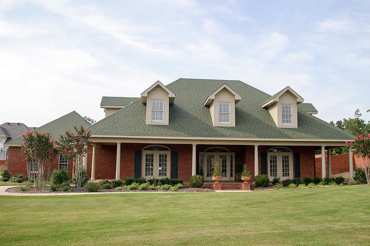 House plan exterior with two stories, a covered porch, multiple dormers, and a green shingle roof. Brick exterior with shutters.