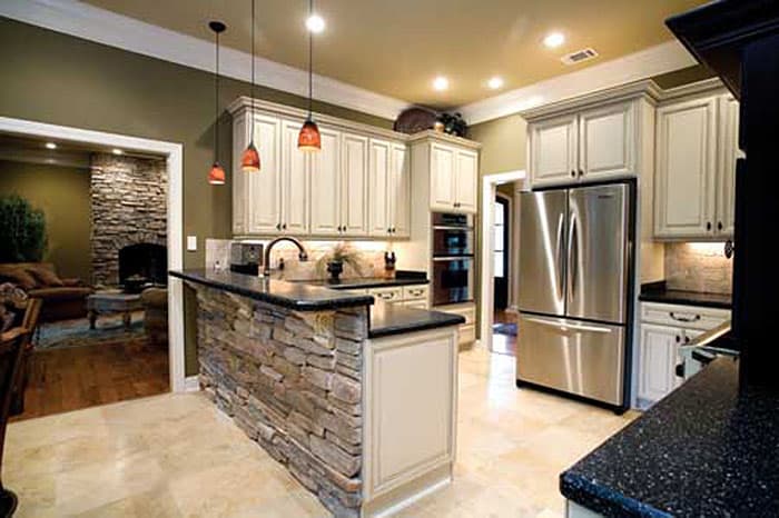 Kitchen with stone-faced island, cream cabinets, stainless steel appliances, and view into living room with stone fireplace.