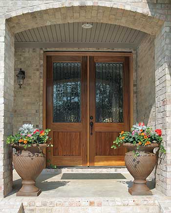 House plan exterior featuring double wooden doors with glass panels under a brick arch and two large urns with flowers.