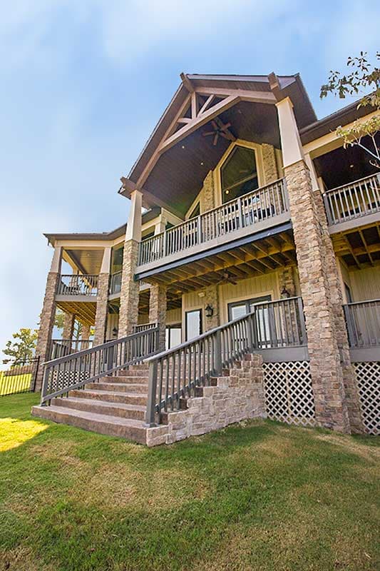 House plan exterior: Stone-clad multi-story home with multiple covered balconies, large gabled dormer, and expansive stone staircase.