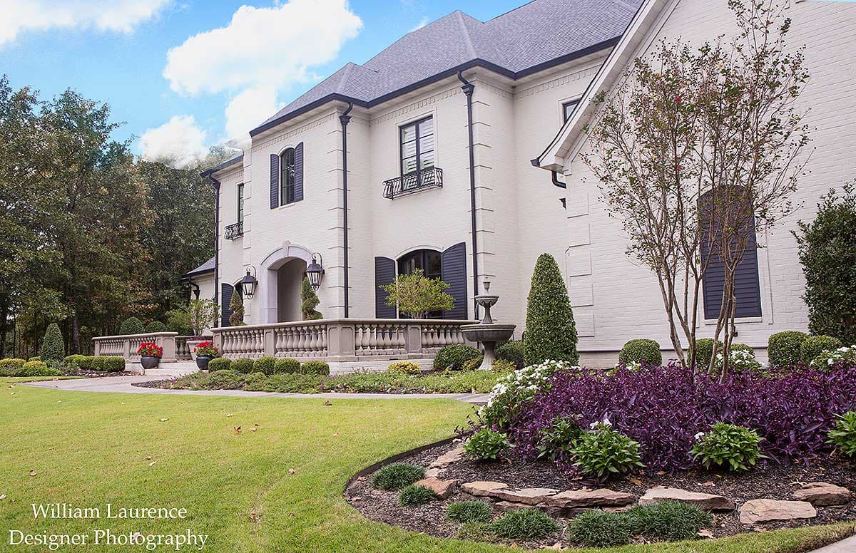 House plan exterior: White brick two-story home with arched entryway, dark shutters, formal balustrade, and landscaping.