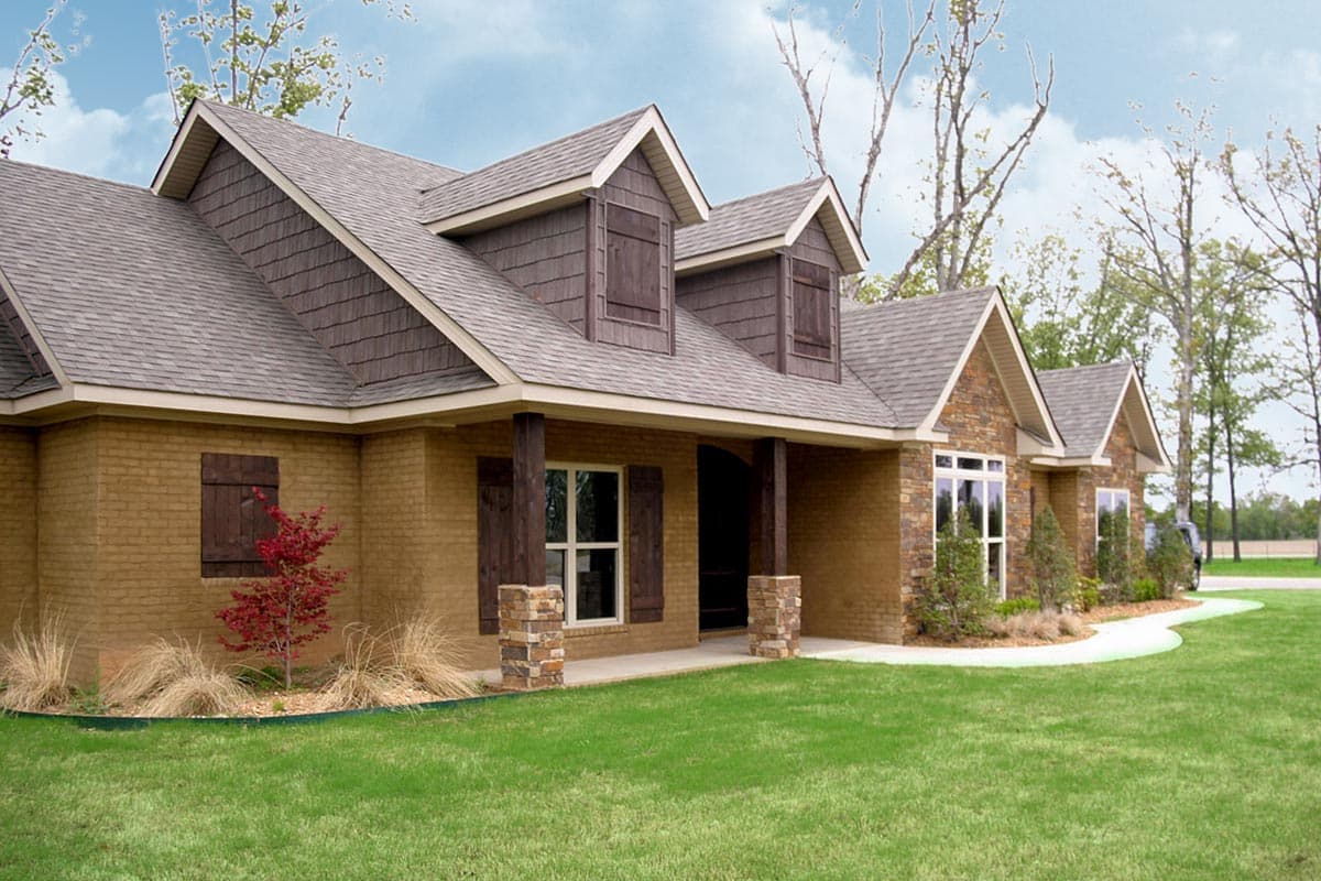House plan exterior with stone accents, dormers with shutters, and covered entry porch supported by stone-wrapped columns.