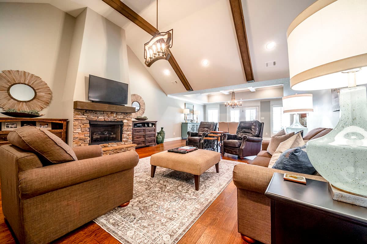 Living room with vaulted ceiling, exposed beams, stone fireplace, seating area, and an open layout to dining and kitchen.