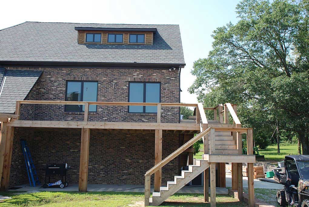 House plan exterior with brick facade, multiple windows, dormers, and a multi-level wooden deck with stairs.