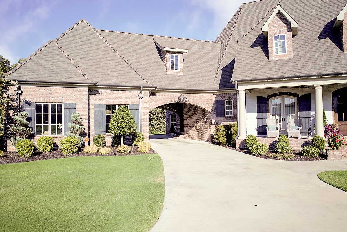 Brick house plan exterior with arched porte-cochère, dormer windows, covered porch with columns, and multi-level rooflines.