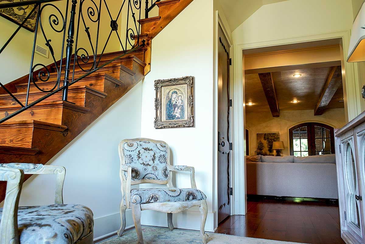 Interior view of a staircase with ornate wrought iron railing, decorative chair, and doorway to a room with exposed wooden beams.