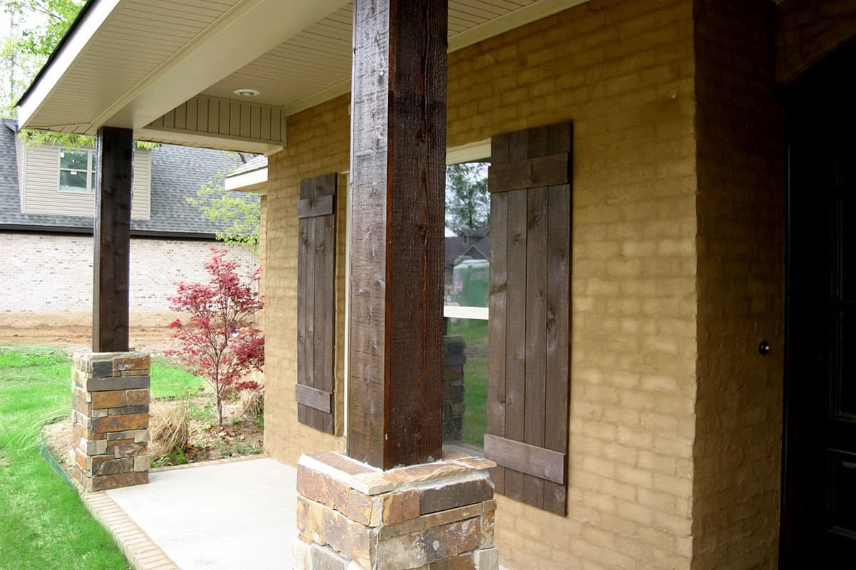 House plan exterior with a covered porch featuring stone-clad columns, wood posts, and brick walls with wood shutters.