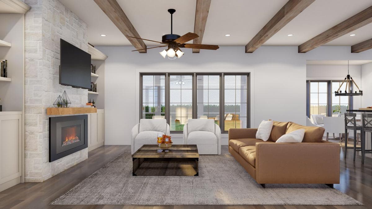 Interior view of a great room with a stone fireplace, vaulted beamed ceiling, and large windows overlooking a yard.