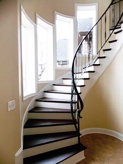 Curved staircase with dark wood treads, white risers, and ornate black railing, illuminated by curved windows.