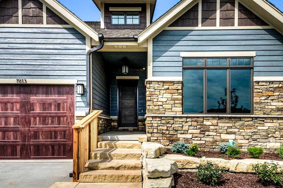 House plan exterior shows blue siding, stone accents, a wooden garage door, and a covered front entrance with stairs.