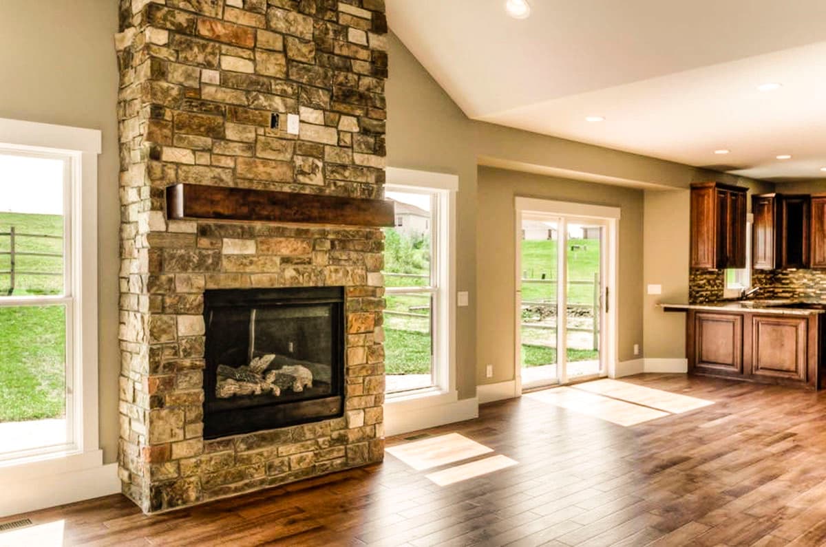 Living area with vaulted ceiling, stone fireplace with wood mantel, windows, and view of kitchen with wood cabinetry.