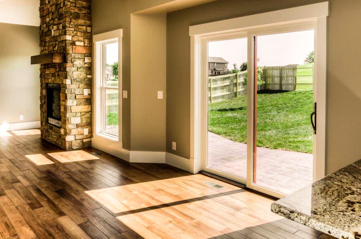 Interior view of a living room featuring a stone fireplace, wood floors, and a sliding glass door opening to a backyard patio and lawn.