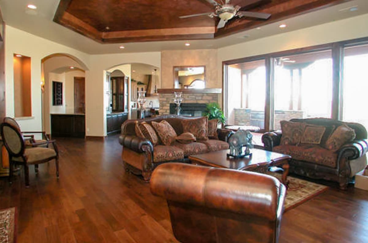 Great room with coffered ceiling, stone fireplace, wet bar, and large windows. Open to kitchen and dining area.