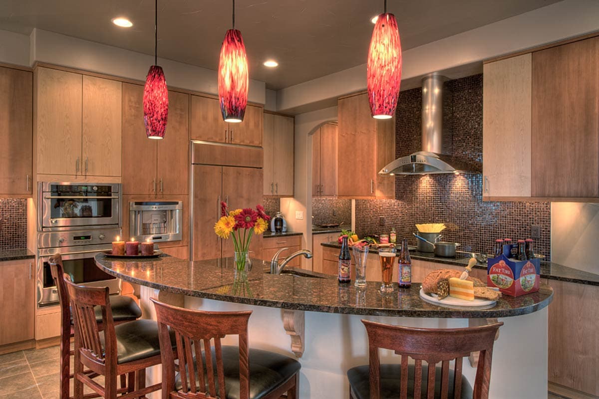 Kitchen with curved granite island, wood cabinetry, stainless steel appliances, pendant lights, and mosaic tile backsplash.