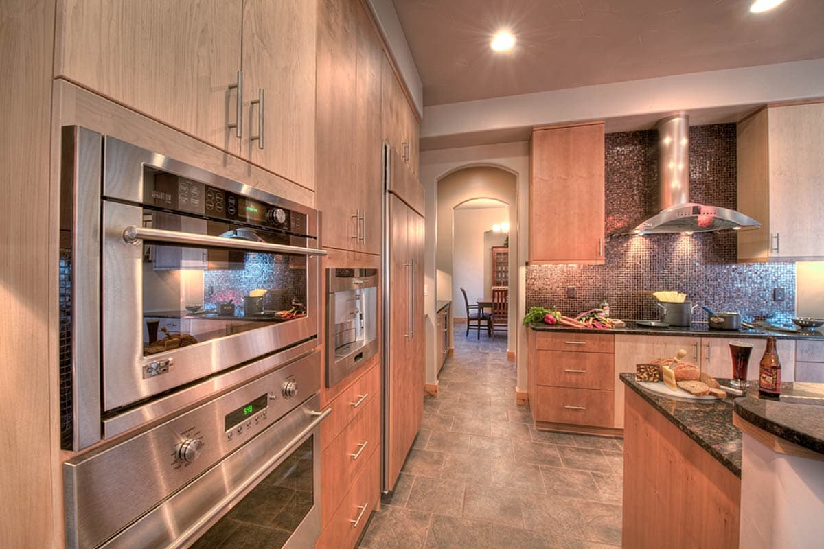 Kitchen interior with stainless steel appliances, wood cabinetry, mosaic tile backsplash, and view of dining area through arched doorway.