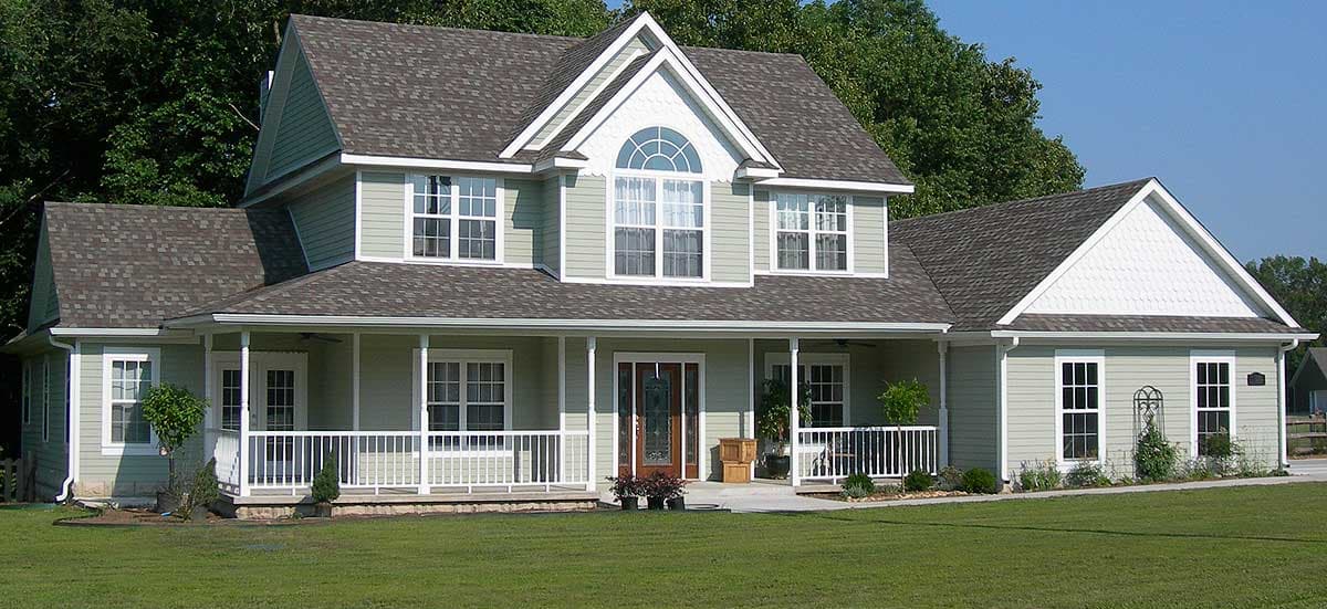 House plan exterior: two-story, light green siding, wraparound porch with white railings, prominent gables, and arched dormer.