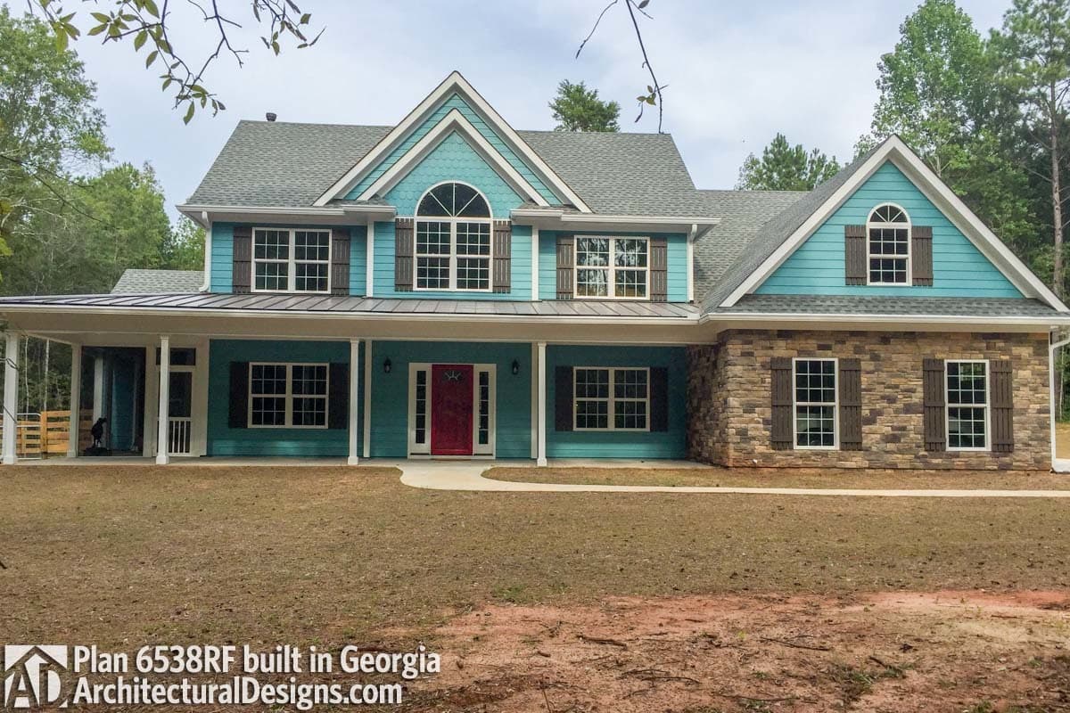 House plan exterior: Two-story home with wraparound porch, stone base, gables, and arched dormer.