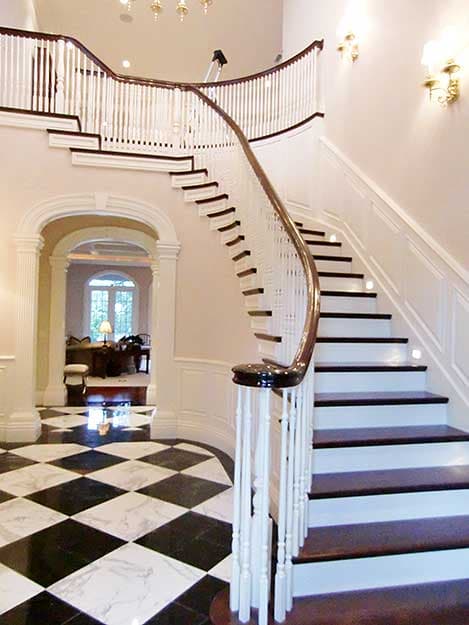 Interior view of a grand foyer with a curved staircase, white balusters, dark wood handrail, and black and white marble floor.