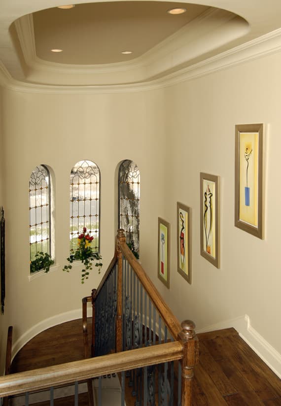 Interior view of a curved staircase with arched windows featuring decorative glass and metalwork, leading to a landing with artwork.