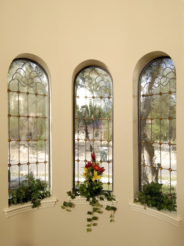 Three tall arched stained glass windows with amber accents and decorative leadwork, flanked by plants.