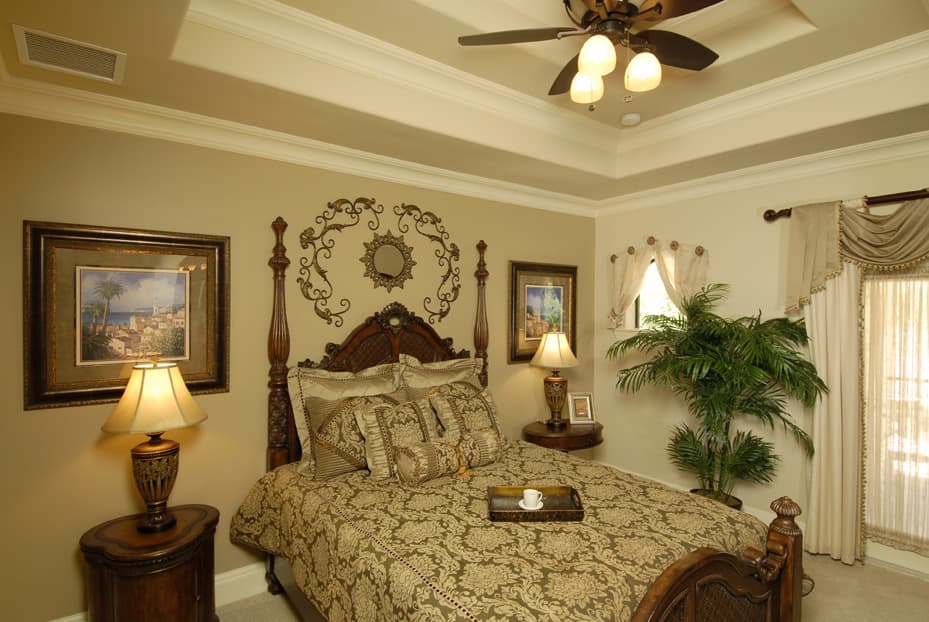 Bedroom with ornate four-poster bed, patterned bedding, tray on bed, two nightstands, framed art, and recessed ceiling with fan.