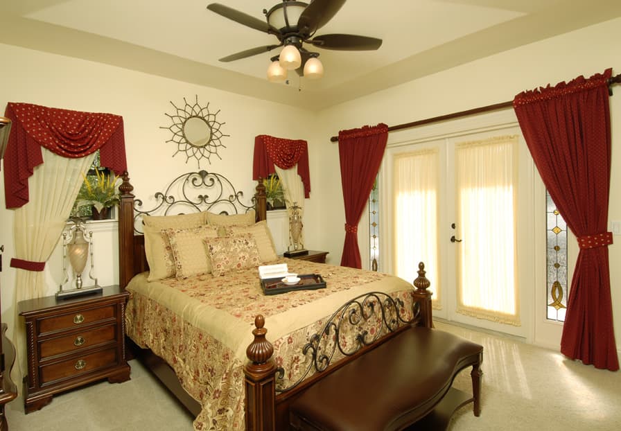 Bedroom interior with ornately decorated metal headboard, patterned bedding, dark wood furniture, and double doors with stained glass sidelights.