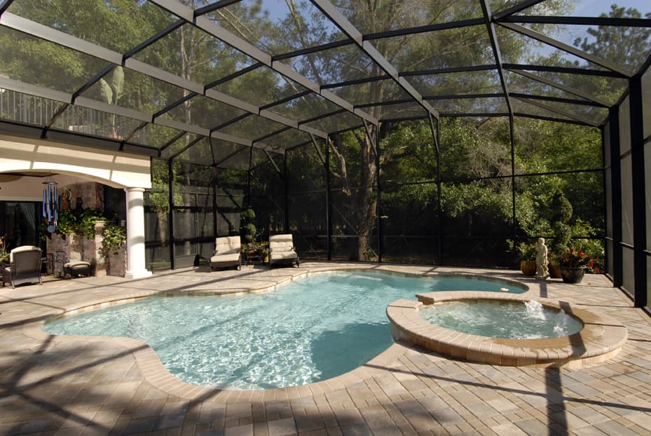 Enclosed pool and spa with brick paver deck, covered patio with columns, and tropical landscaping.