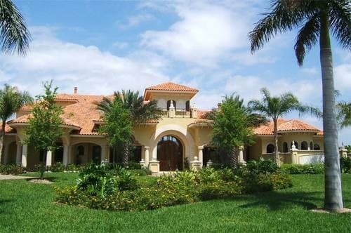 House plan exterior: Large Mediterranean-style home with red tile roof, arched entryway, multiple gables, and palm trees.