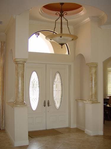 Interior view of a grand entryway featuring double doors with oval stained-glass panels, decorative columns, and a recessed ceiling.