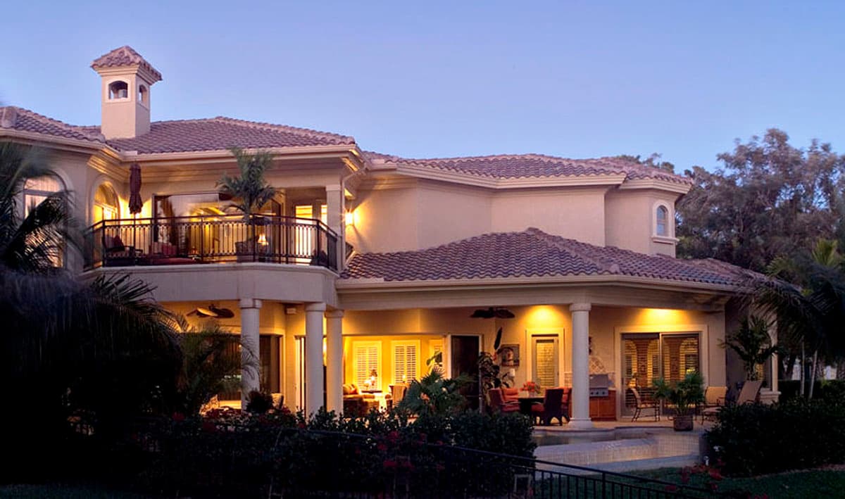 House plan exterior with a prominent balcony, arched windows, and covered patio areas with outdoor dining. Two stories visible.