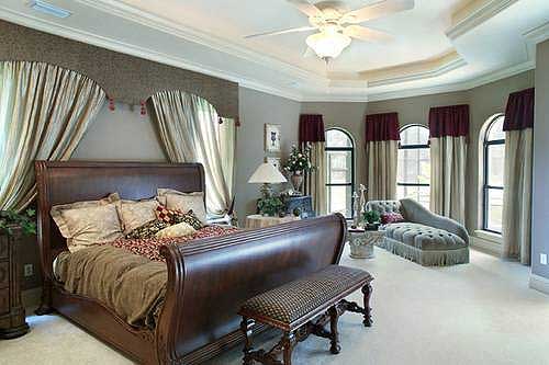 Primary bedroom with sleigh bed, arched windows with draped curtains, and a tray ceiling with a ceiling fan.
