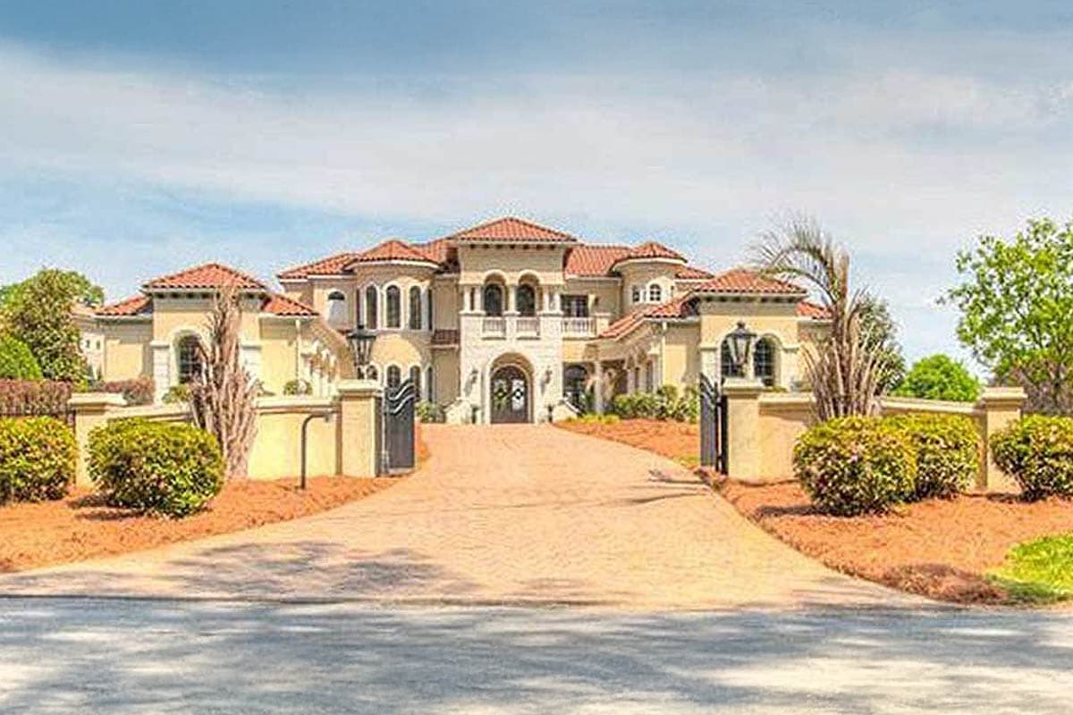 House plan exterior. Two-story Mediterranean-style home with arched windows and entryway, a red tile roof, and a gated driveway.
