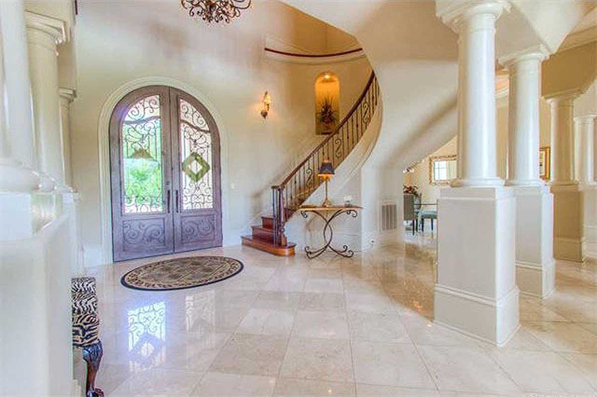 Grand foyer with a curved staircase, ornate double doors with wrought iron details, marble flooring, and classical columns.