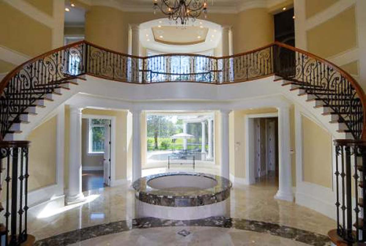 Interior view of a grand foyer with a curved double staircase, marble flooring, and a circular central feature.
