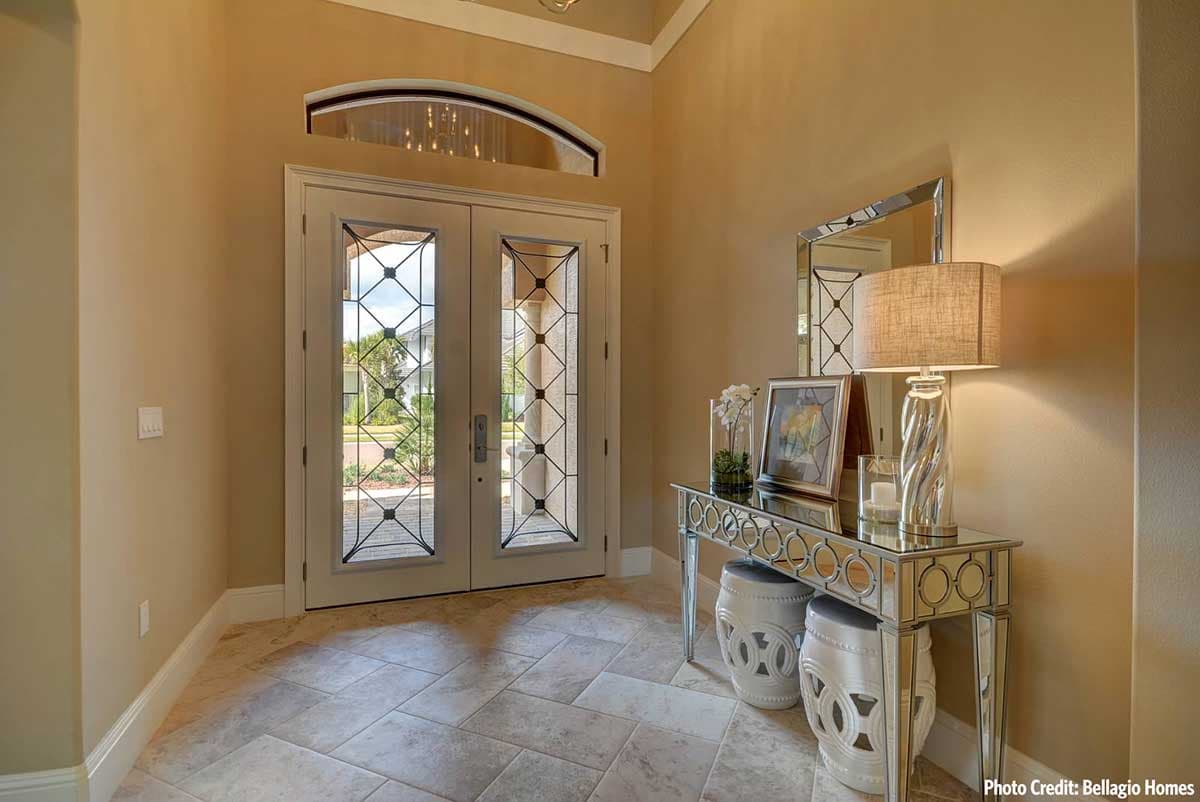 Foyer interior with double doors featuring decorative glass panes, arched transom window, mirrored console table, and stone tile flooring.