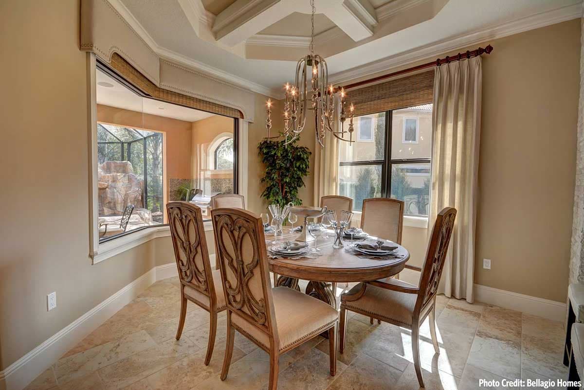 Dining room with round table, upholstered chairs, coffered ceiling, and large bay window overlooking a pool area.