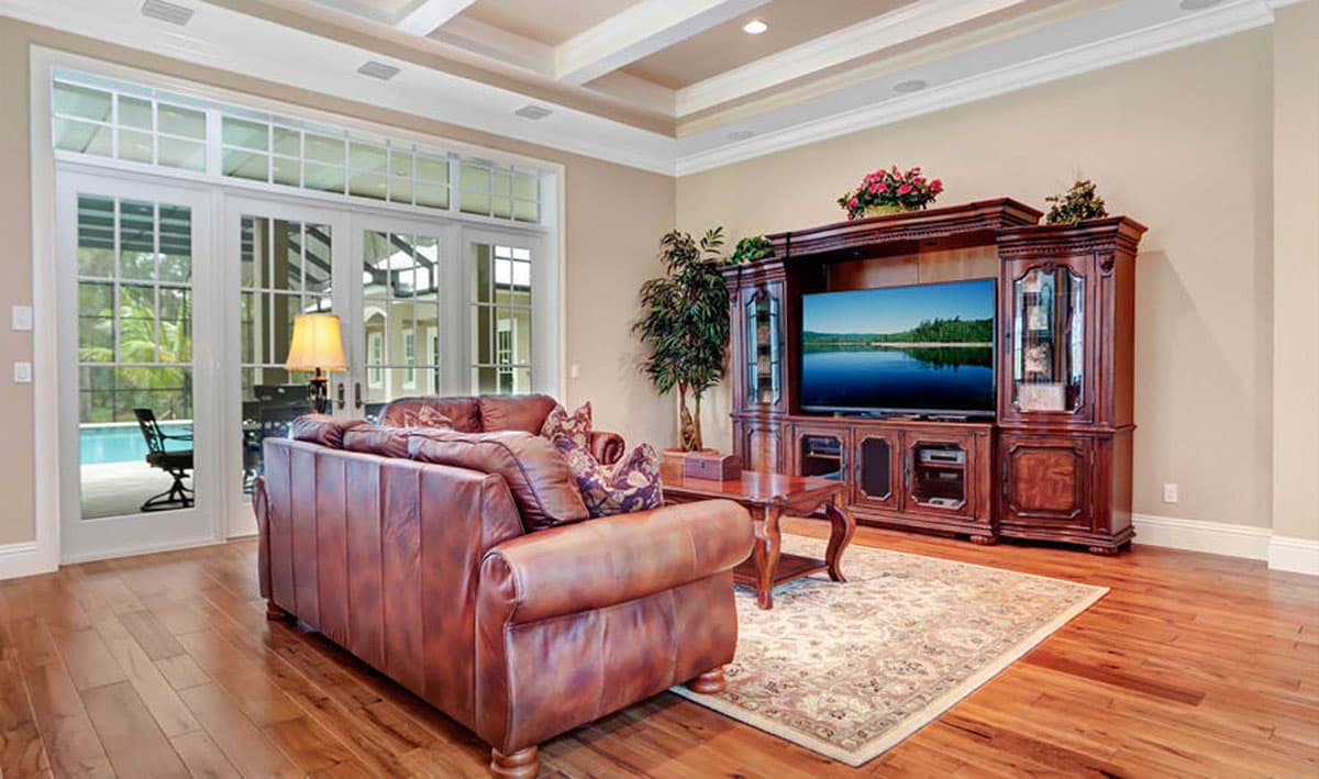 Living room interior with coffered ceiling, leather sofa, ornate media console, and french doors opening to a patio and pool.