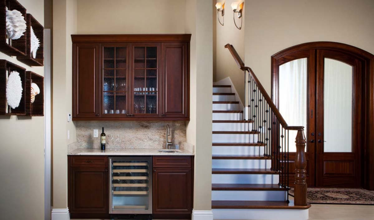 Interior view of a home entrance with a wooden staircase, dark wood cabinets, wine fridge, and double doors.