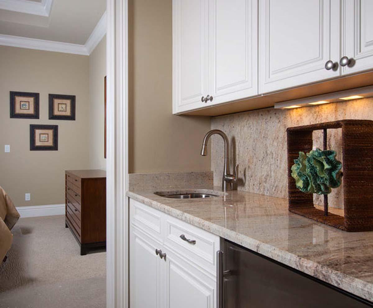 Wet bar with white cabinetry, granite countertop, stainless steel sink, and faucet. Adjacent room shows framed art.