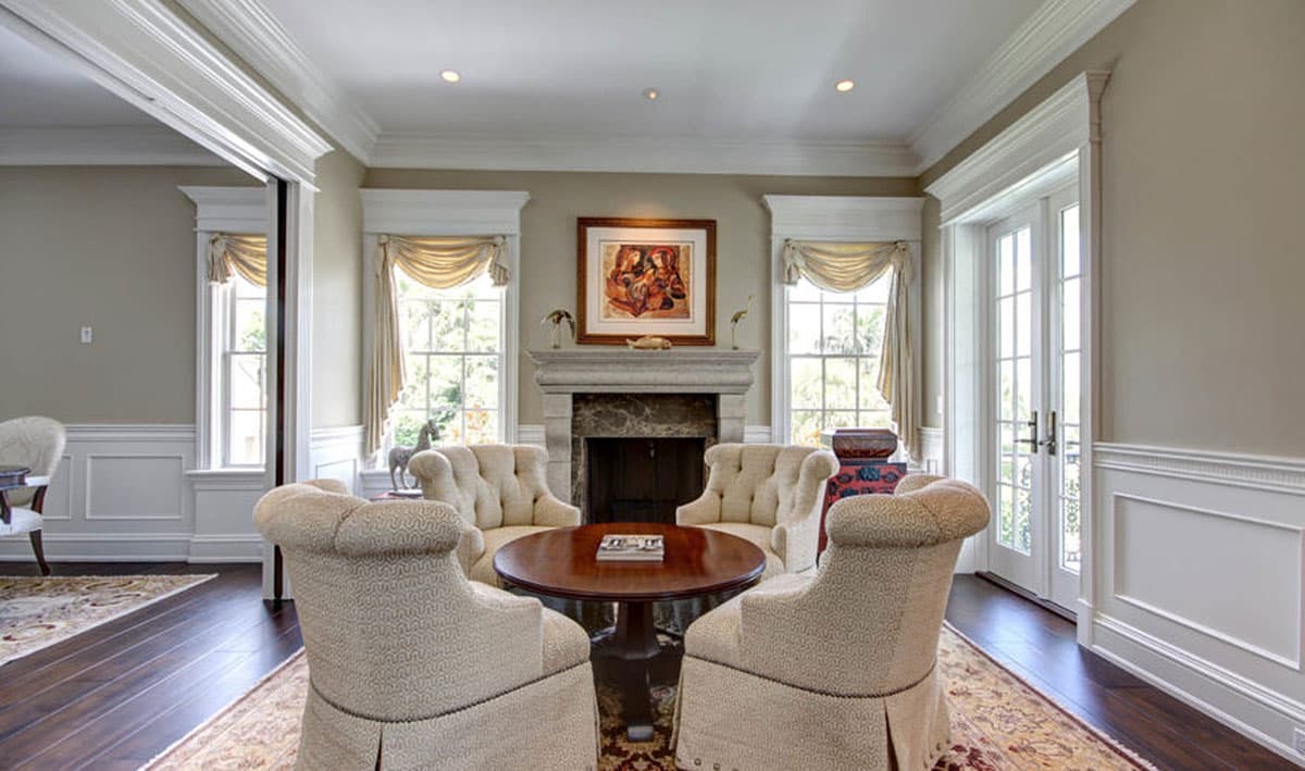 Interior view of a living room with fireplace, seating area, and French doors. Detailed molding and paneled walls are visible.