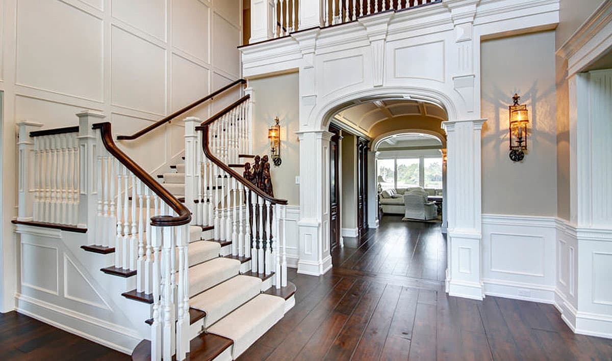 Interior view of a grand foyer with a curved staircase, paneled walls, arched doorway, and dark wood flooring.