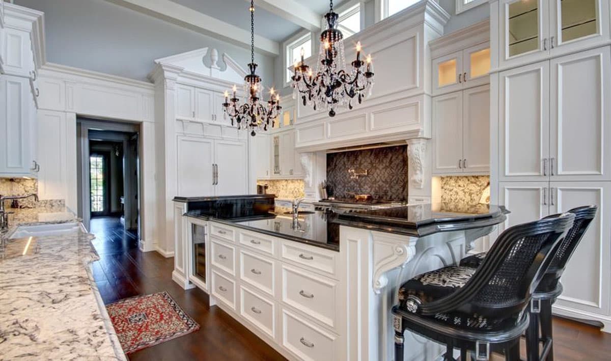 Spacious kitchen with large island, granite countertops, ornate cabinetry, dual chandeliers, and an open doorway to another room.