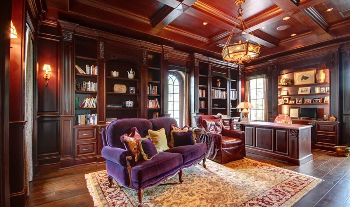 Library with extensive dark wood built-in bookshelves, a large desk, purple sofa, leather armchair, and ornate rug.