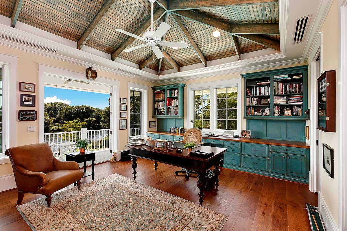 Home office with vaulted wood-beamed ceiling, built-in teal cabinetry, desk, leather armchair, and French doors to balcony.