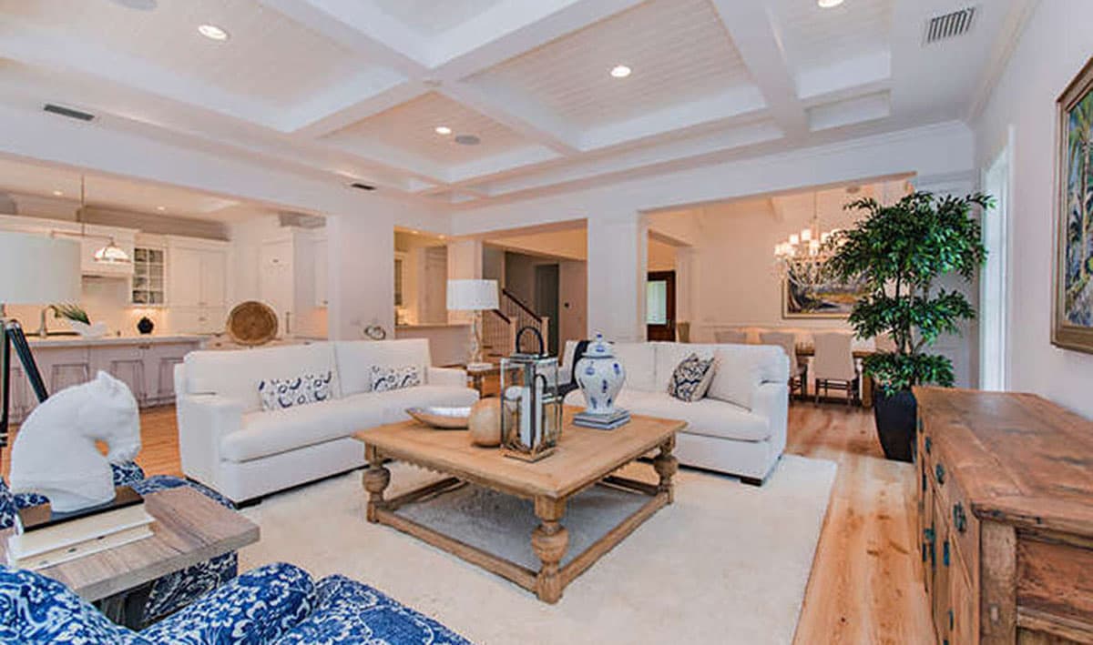 Living room with coffered ceiling, white sofas, coffee table, and view of kitchen and dining area.