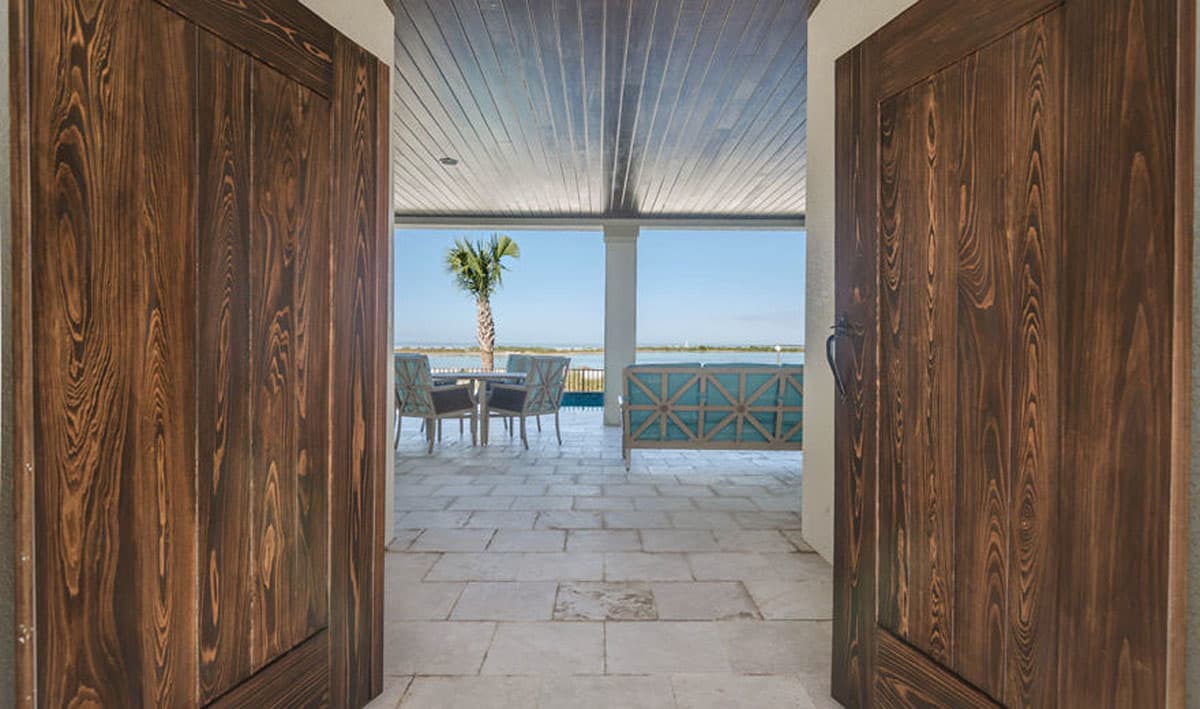 View of covered patio with seating and dining sets through double wooden doors with lake view.