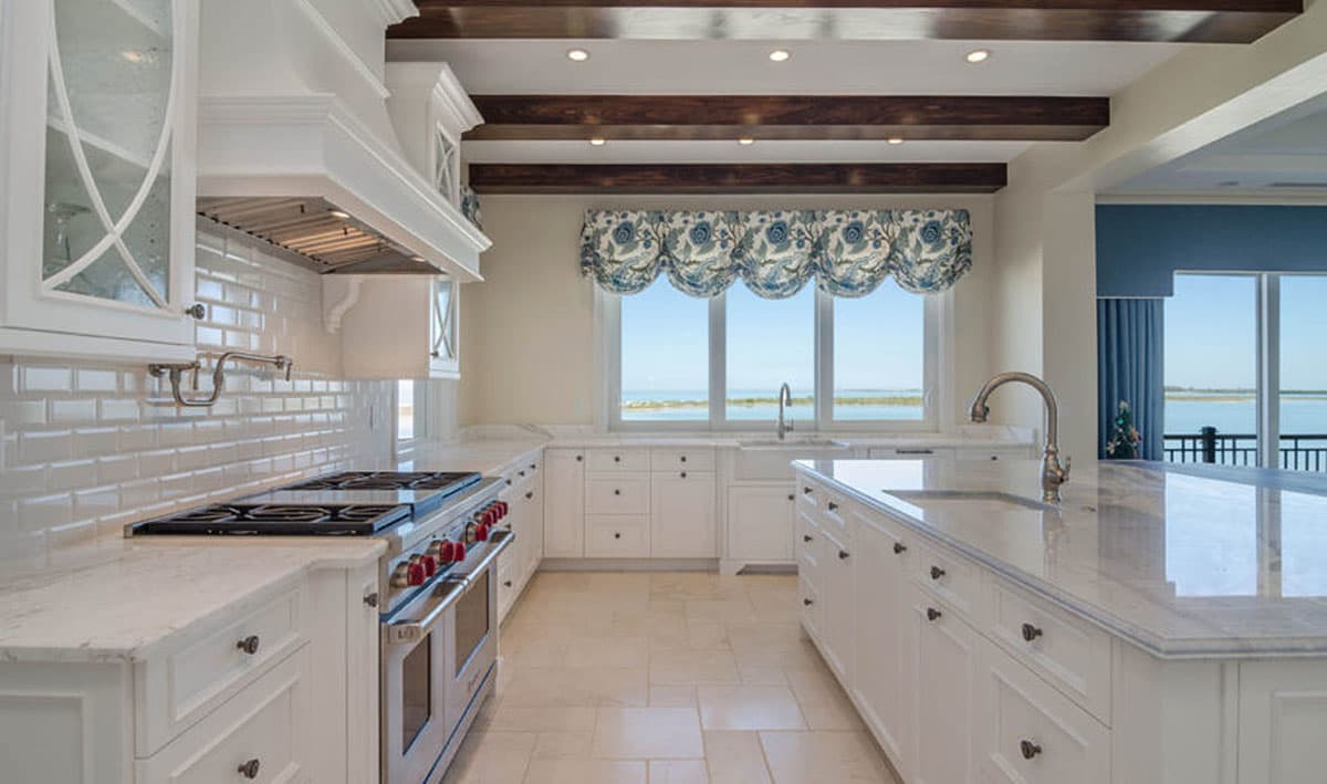 Interior view of a kitchen with white cabinetry, subway tile backsplash, large range, island, and window overlooking water.